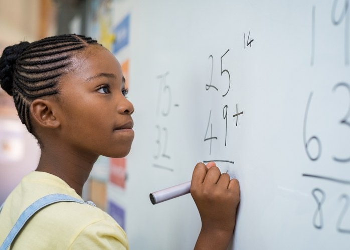 Portrait of african girl writing solution of sums on white board at school. Black schoolgirl solving addition sum on white board with marker pen. School child thinking while doing mathematics problem.