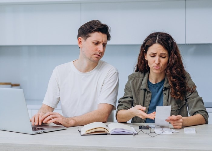Worried married couple going through bills and managing finances at home using laptop and calculator, looking at store receipts, receipts and documents, faces budgeting problems.